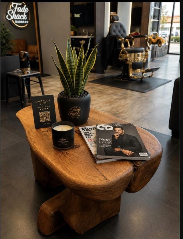 Inside Fade Shack Barbers, Orewa: a live-edge wooden table with a Fade Shack plant pot, candle and magazines, and an ornate gold barber chair behind.