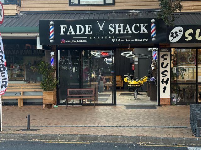 The Fade Shack Barbers shopfront on Moana Avenue, Orewa — black signage with barber poles and scissors, the Open sign lit, and gold barber chairs through the glass.